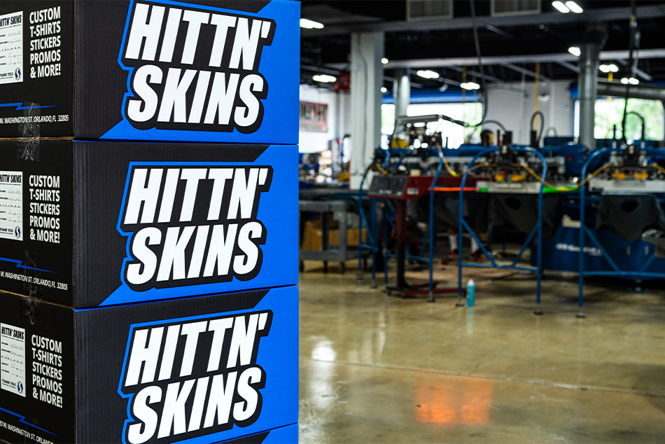 Hittn' Skins Packaging Boxes Stacked with M&R Screen Printing Automatic Press Machine in the Background