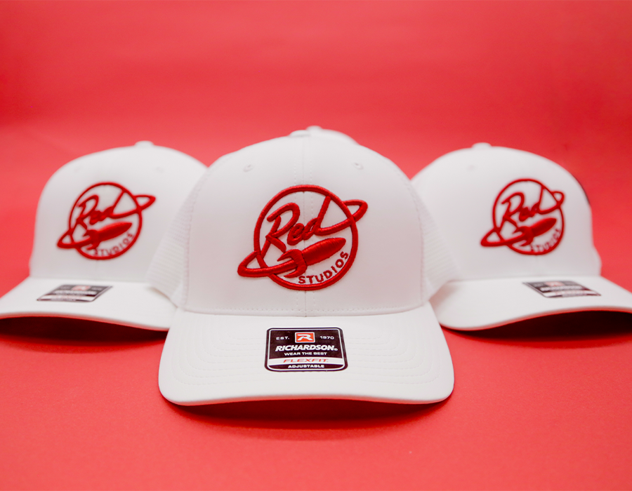 Three white Richardson hats featuring custom red embroidered Red Studios logos displayed against a bright red background.