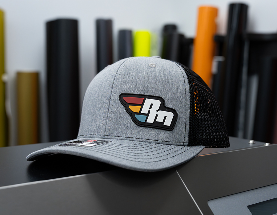 Heather gray and black custom trucker hat featuring a multi-color molded PVC logo patch on the front.