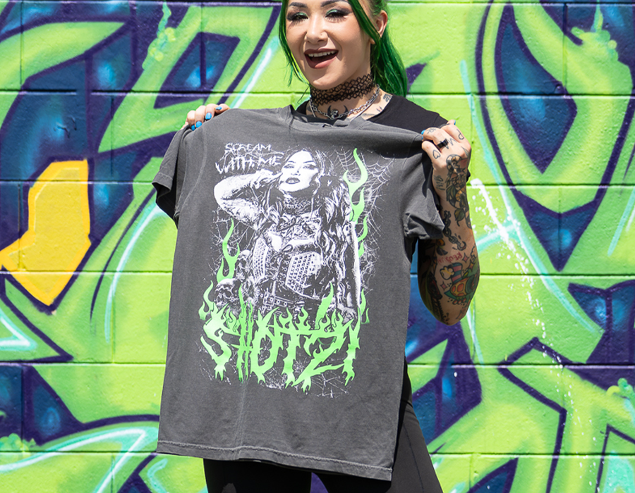 Shotzi modeling a charcoal custom screen printed wrestling T shirt featuring large black, white and green illustrated artwork in front of a graffiti wall.