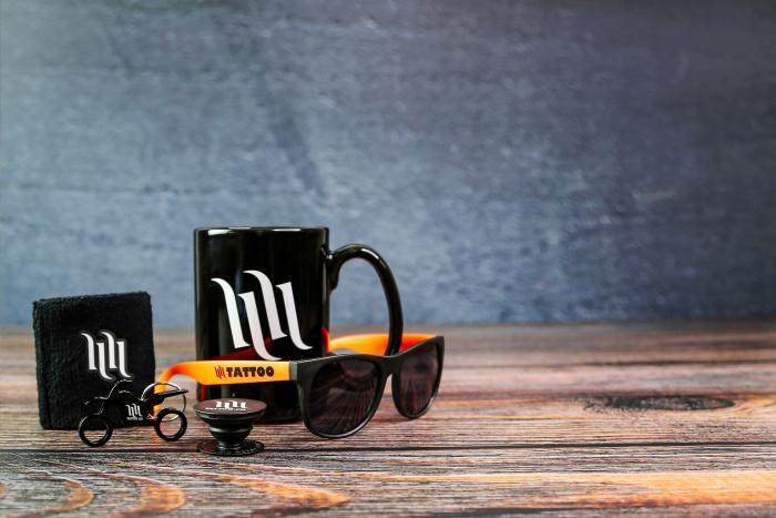 Branded promotional merchandise set featuring a black mug, sunglasses with orange arms, wristband, pop socket, and a custom metal keychain in the shape of a bicycle—all displayed on a rustic wooden surface with a dark textured backdrop.